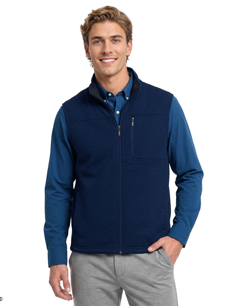 A man wearing a navy blue zip-up fleece vest over a blue button-down shirt and gray pants, standing against a plain white background.