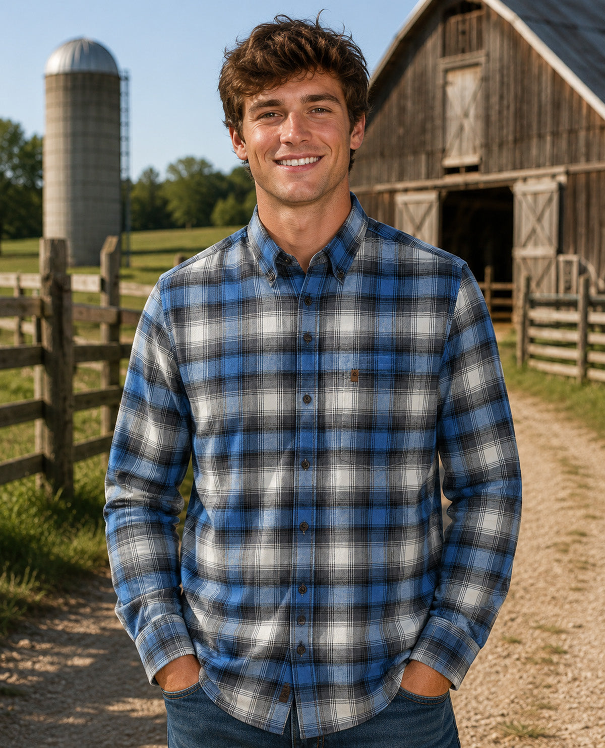 A smiling young man stands outdoors on a farm path wearing a blue, gray, and white plaid button-down flannel shirt, with a barn, silo, and fence in the background.