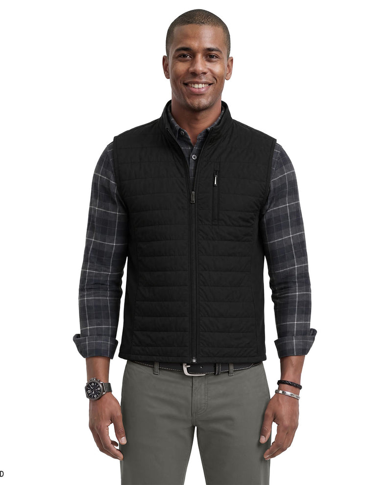 A man wearing a black quilted zip-front vest over a dark gray plaid button-down shirt, paired with gray trousers, against a plain white background.