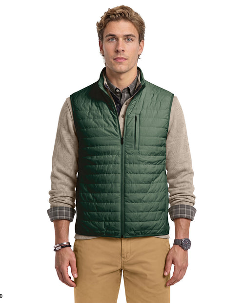 A male model wearing a dark green quilted puffer vest over a beige and gray knit sweater, plaid shirt, and tan pants, photographed against a plain white background.