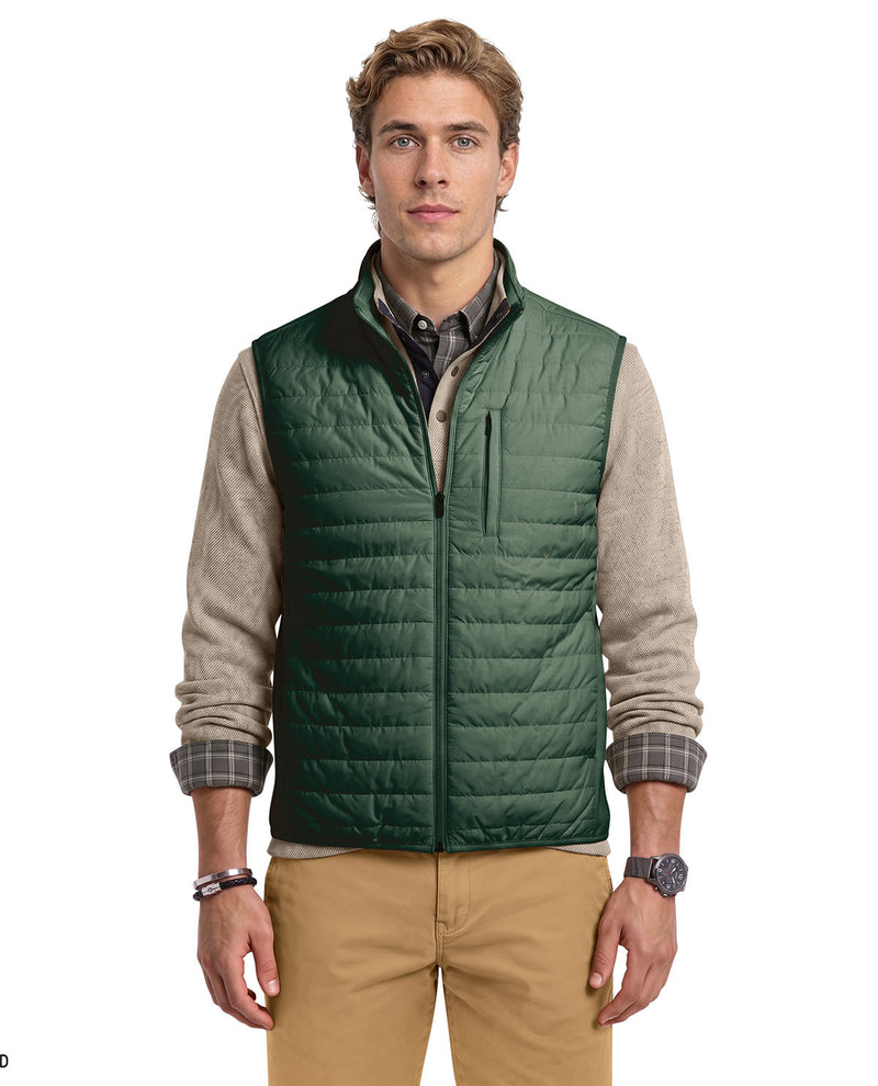 A male model wearing a dark green quilted puffer vest over a beige and gray knit sweater, plaid shirt, and tan pants, photographed against a plain white background.