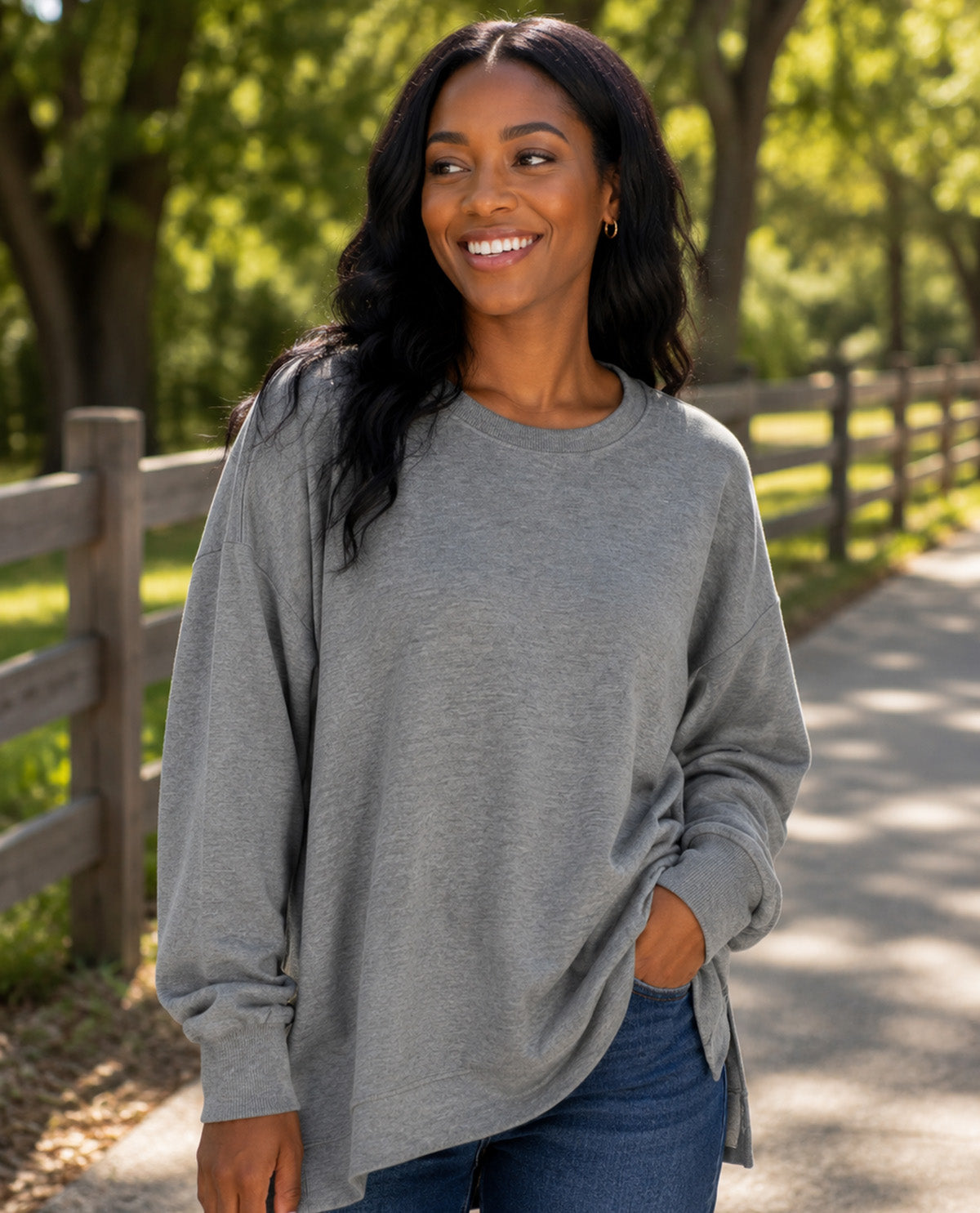 A woman wearing a heather gray long-sleeve crewneck sweatshirt and blue jeans, standing on a paved path in a park with trees and a wooden fence in the background.