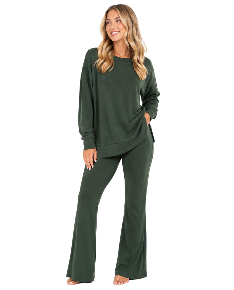 A woman modeling a dark green two-piece loungewear set consisting of a long-sleeve crewneck top and matching wide-leg pants, set against a plain white background.