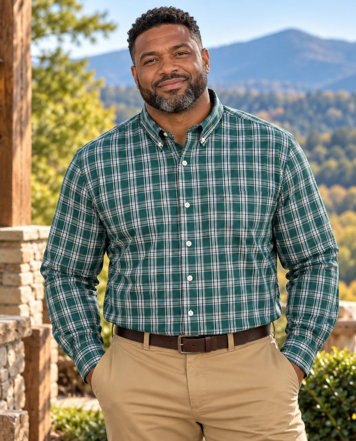 A man wearing a green and white plaid long-sleeve button-down shirt with khaki pants, standing outdoors in front of a scenic mountain background.