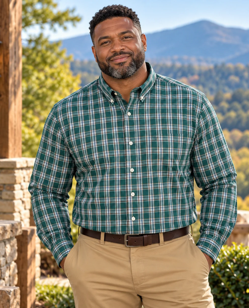 A man wearing a green and white plaid long-sleeve button-down shirt with khaki pants, standing outdoors in front of a scenic mountain background.