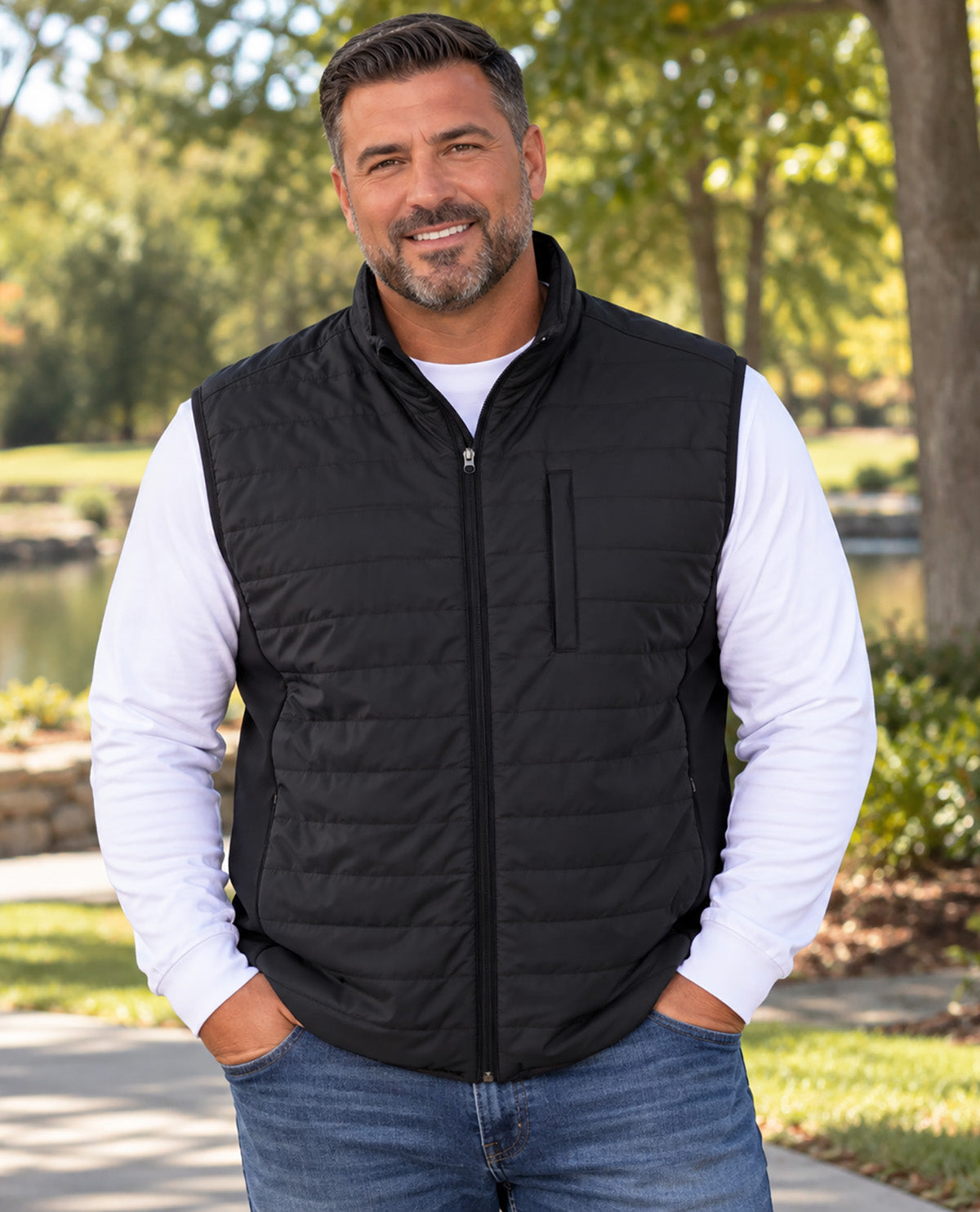 A smiling man wearing a black zip-up quilted vest over a white long-sleeve shirt and blue jeans, standing outdoors on a paved path with trees and grass in the background.