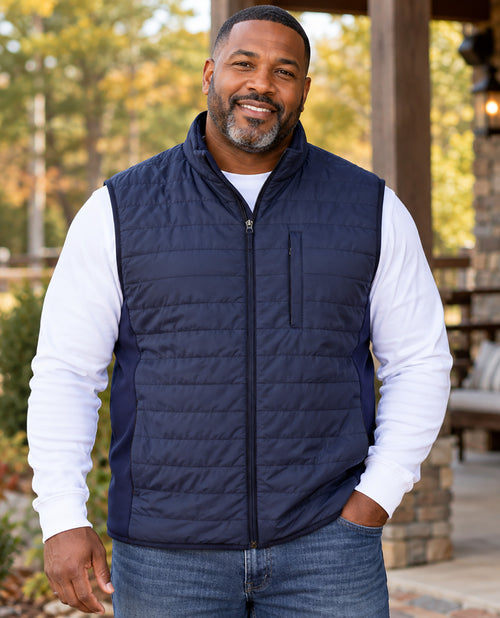 A smiling man wearing a navy blue quilted zip-up puffer vest over a white long-sleeve shirt and blue jeans, standing outdoors in front of a wooden porch with trees in the background.