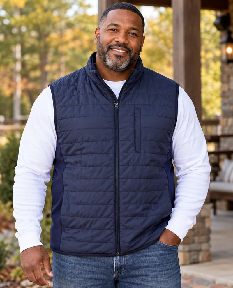 A smiling man wearing a navy blue quilted zip-up puffer vest over a white long-sleeve shirt and blue jeans, standing outdoors in front of a wooden porch with trees in the background.