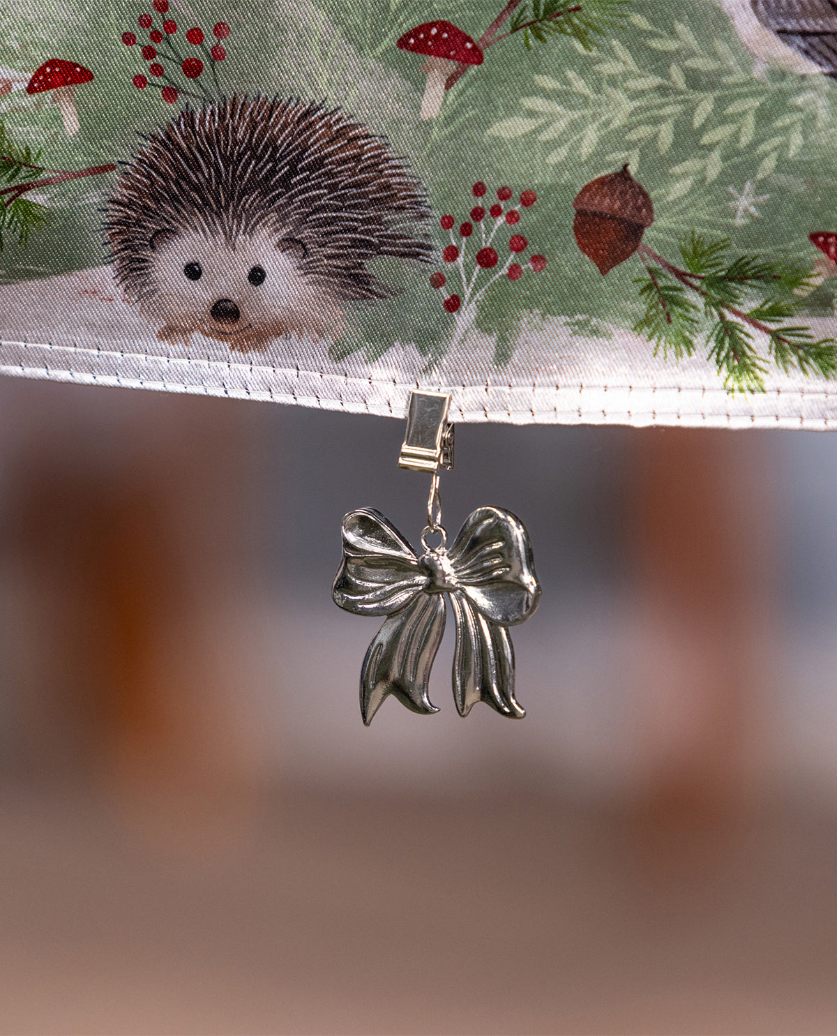 A silver metallic bow-shaped clip or charm hanging from a white stitched edge, with a printed fabric above showing a hedgehog, red mushrooms, berries, and evergreen branches on a soft blurred background.