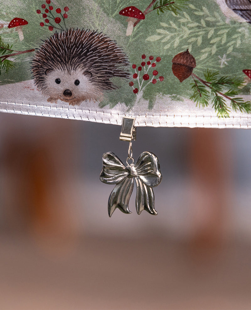 A silver metallic bow-shaped clip or charm hanging from a white stitched edge, with a printed fabric above showing a hedgehog, red mushrooms, berries, and evergreen branches on a soft blurred background.