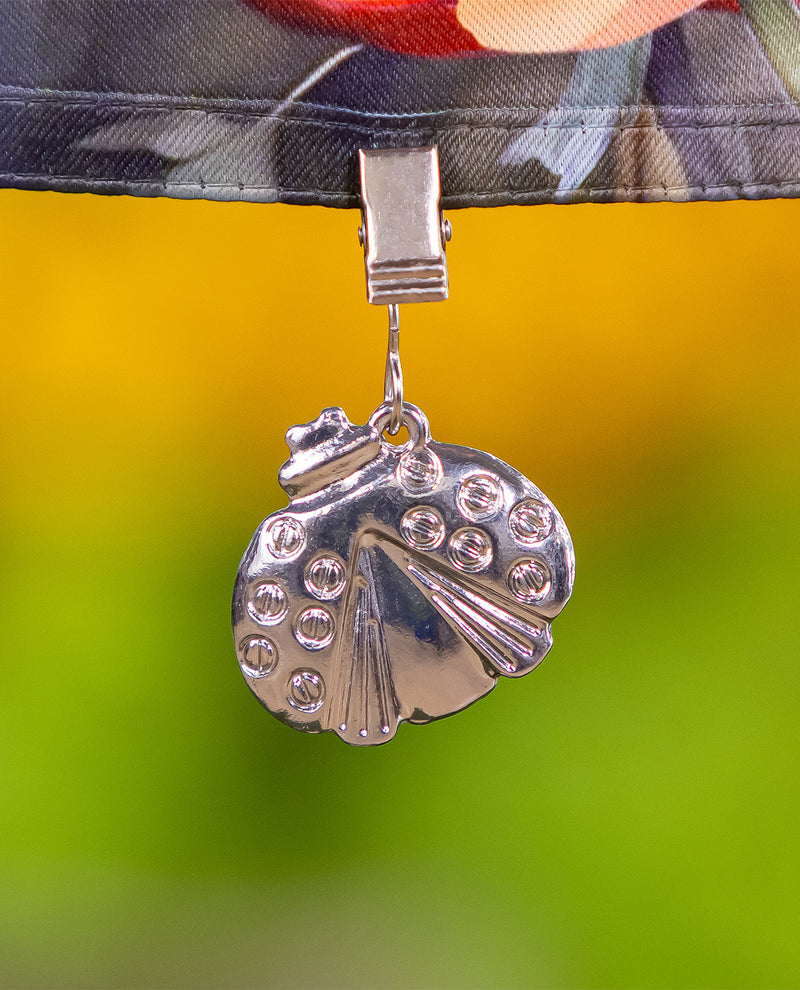 A silver metal peacock-shaped clip or charm hanging from a camouflage-patterned strap, with a bright yellow and green blurred outdoor background.