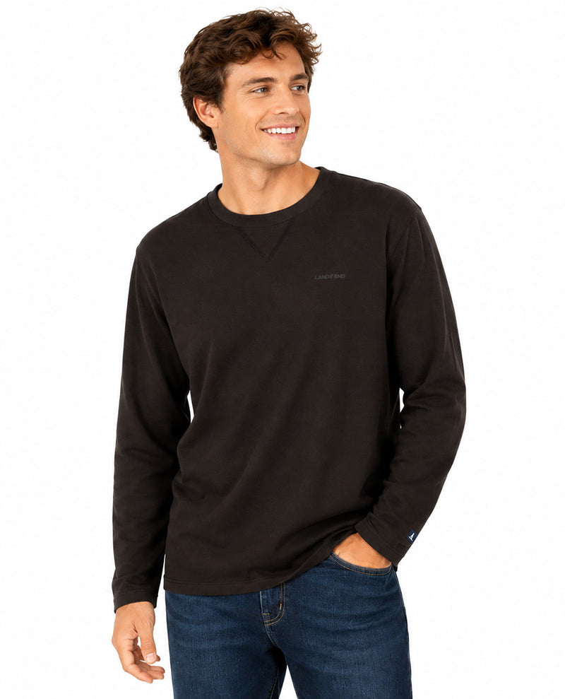 A smiling man wearing a dark brown long-sleeve crewneck shirt and blue jeans, standing against a plain white background.