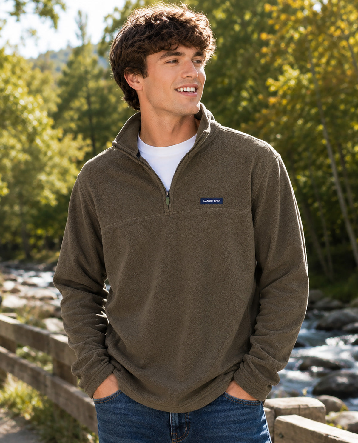 A man wearing a brown quarter-zip fleece pullover by Lands' End, standing outdoors near a wooden fence and a flowing stream with trees in the background.