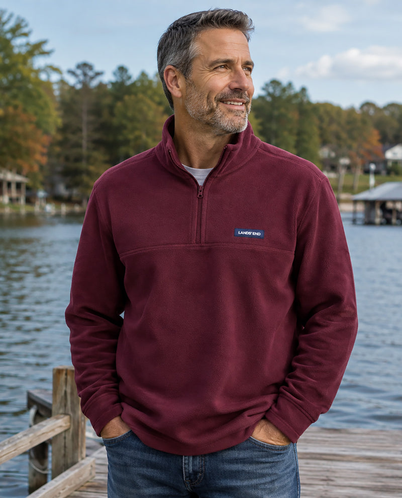 A man wearing a maroon quarter-zip fleece pullover by Landmark stands on a wooden dock with a lake and trees in the background.