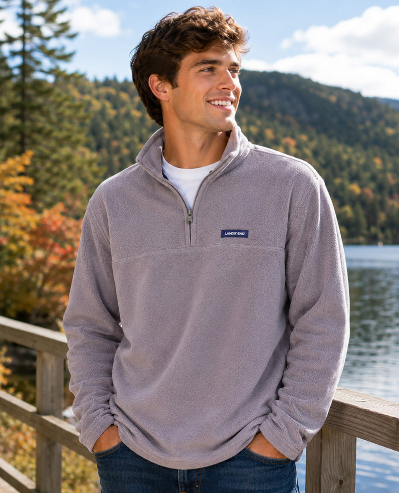 A man wearing a light gray quarter-zip fleece pullover with a small blue and white 'Lands' End logo patch on the chest, standing outdoors by a wooden railing with a lake and forested hills in the background.