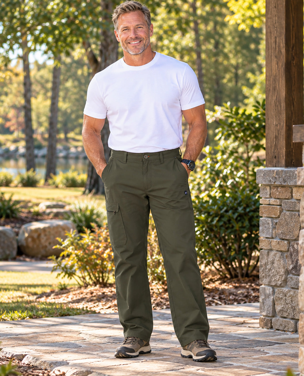 A smiling man standing outdoors on a stone walkway wearing olive green cargo pants, a white t-shirt, and hiking shoes, with trees and landscaping in the background.