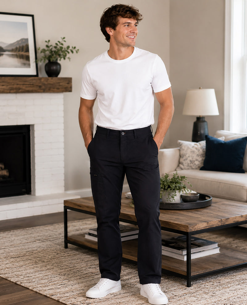 A man wearing a white crewneck t-shirt and dark charcoal gray trousers stands in a modern living room setting with a fireplace, coffee table, and sofa in the background.