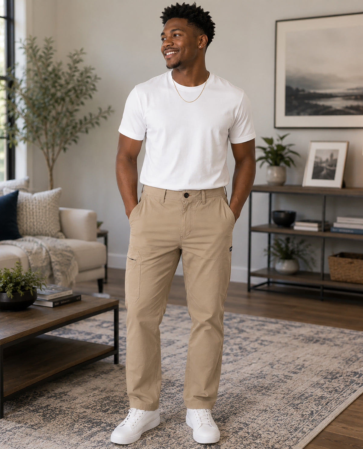 A man wearing beige cargo-style chinos, a white crewneck t-shirt, and white sneakers stands in a modern living room setting.