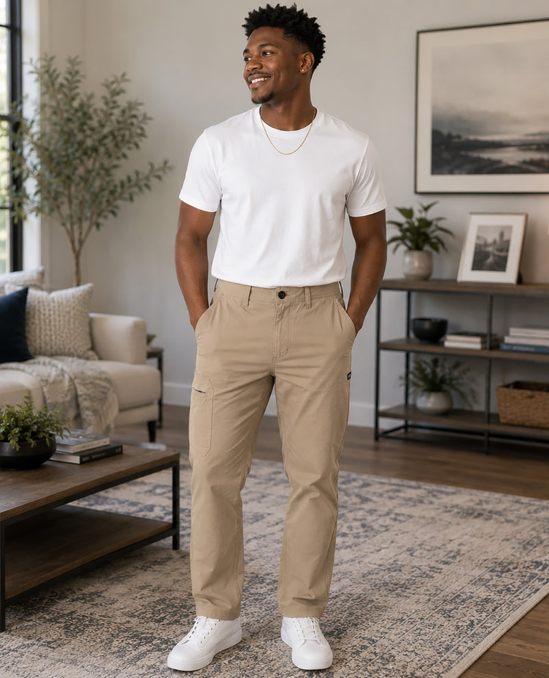 A man wearing beige cargo-style chinos, a white crewneck t-shirt, and white sneakers stands in a modern living room setting.