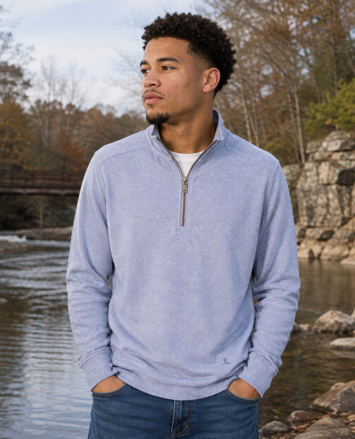A man wearing a light blue heathered quarter-zip pullover sweater and blue jeans, standing outdoors near a river with trees and rocks in the background.