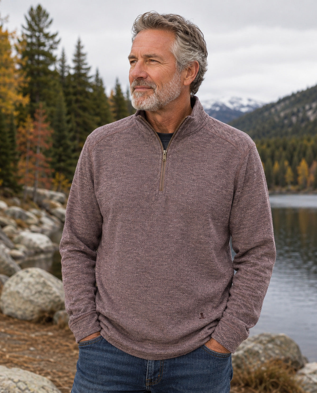 A man wearing a heathered taupe-brown quarter-zip pullover sweater and blue jeans, standing outdoors by a lake with trees and mountains in the background.