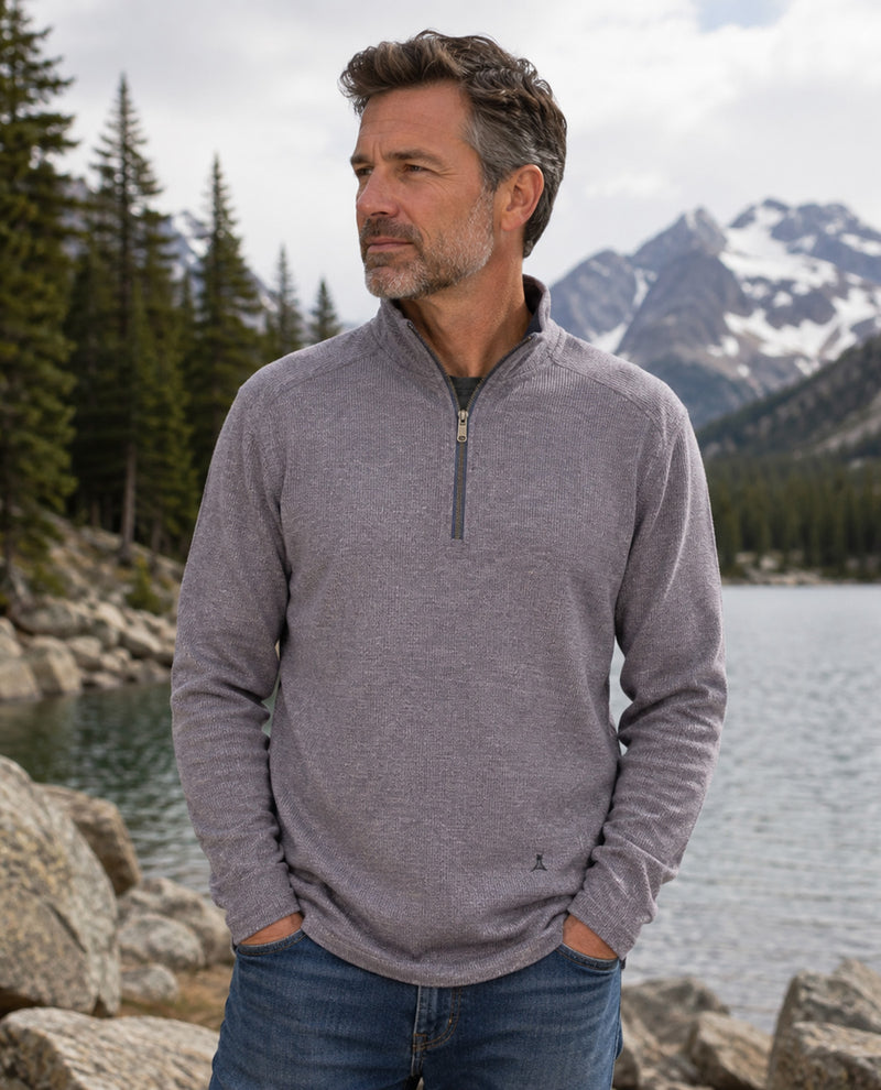 A man wearing a heather gray quarter-zip pullover sweater stands outdoors in front of a lake, mountains, and pine trees, with his hands in his pockets.