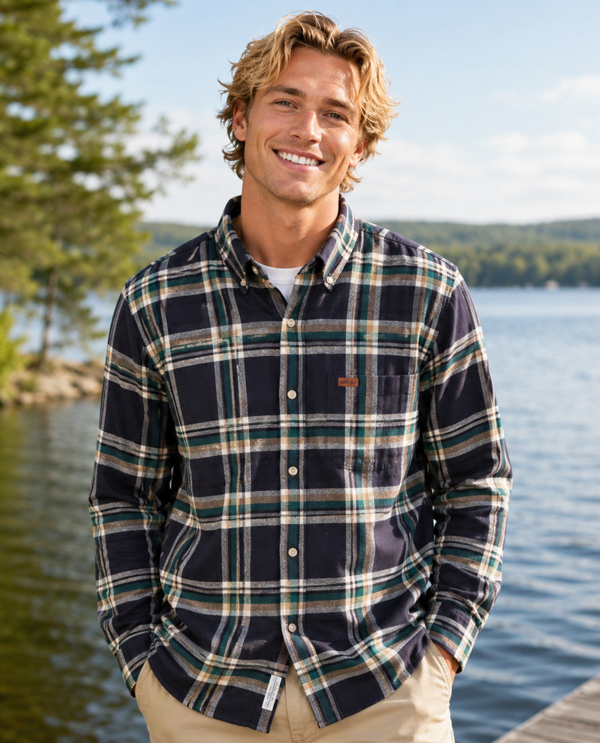 A smiling man standing outdoors by a lake wearing a long-sleeve plaid button-up shirt in navy, green, and beige tones, paired with khaki pants.