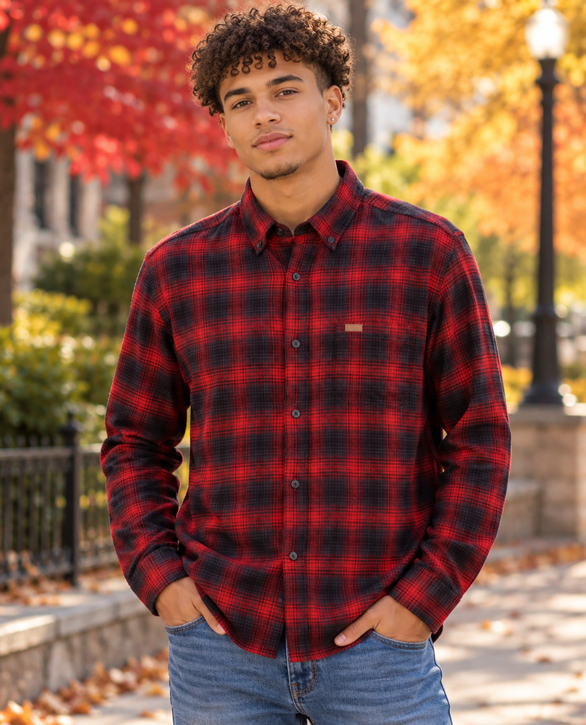 A man wearing a red and black plaid button-down flannel shirt and blue jeans, standing outdoors on a sidewalk with autumn trees in the background.