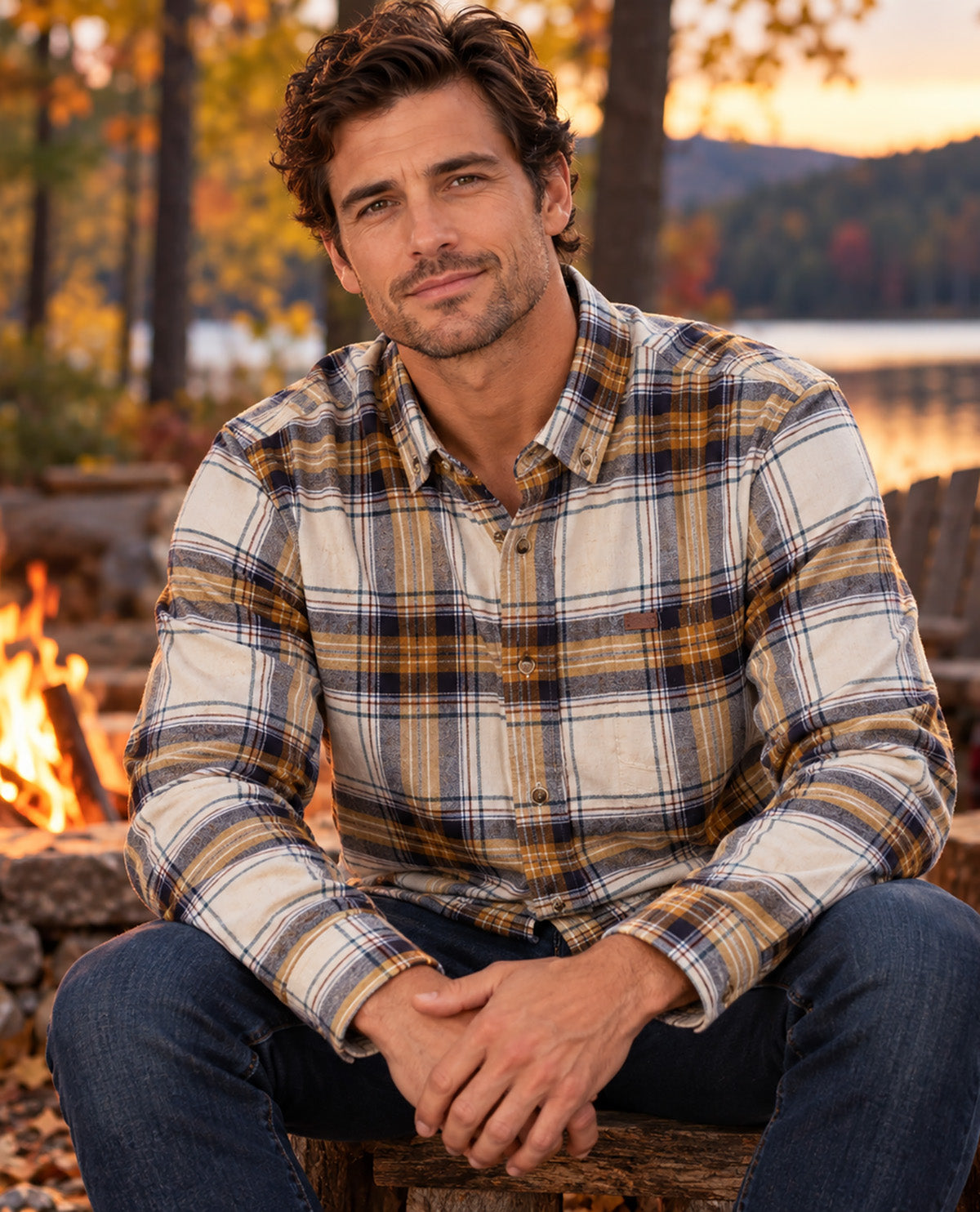 A man sitting outdoors by a campfire at sunset wearing a long-sleeve plaid button-up shirt in cream, brown, and navy, paired with blue jeans.