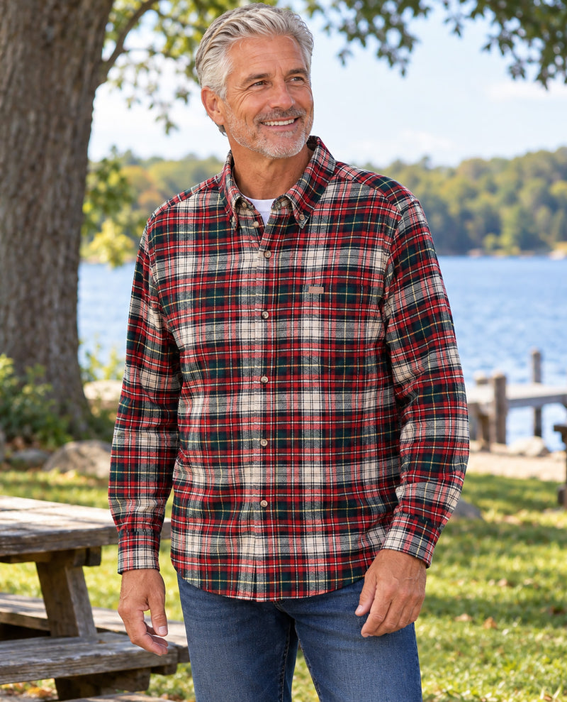 A smiling older man standing outdoors by a lake wearing a long-sleeve plaid button-up shirt with red, green, black, and beige tones, paired with blue jeans.