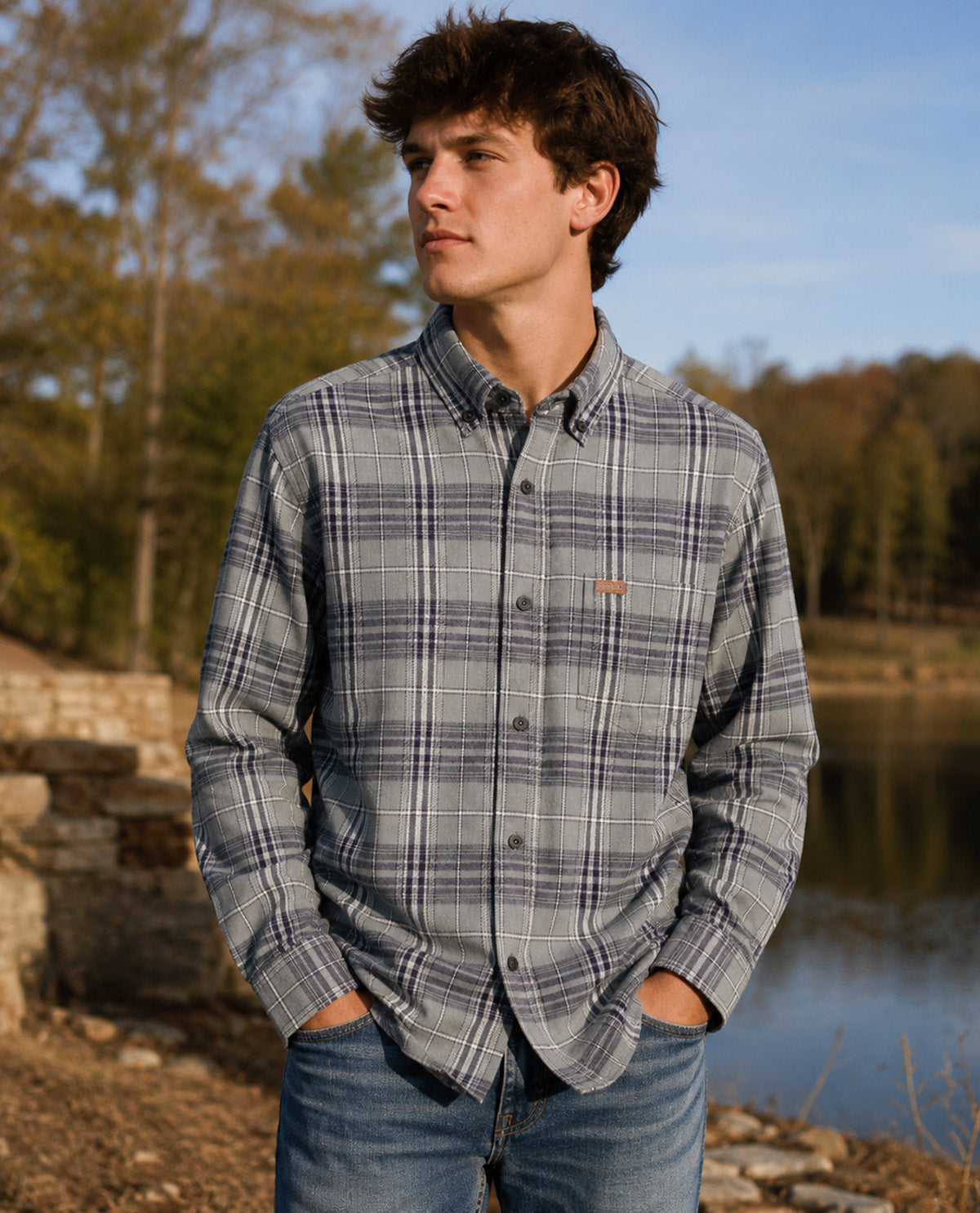 A young man wearing a gray and navy blue plaid button-down shirt with blue jeans, standing outdoors near a lake with trees in the background.