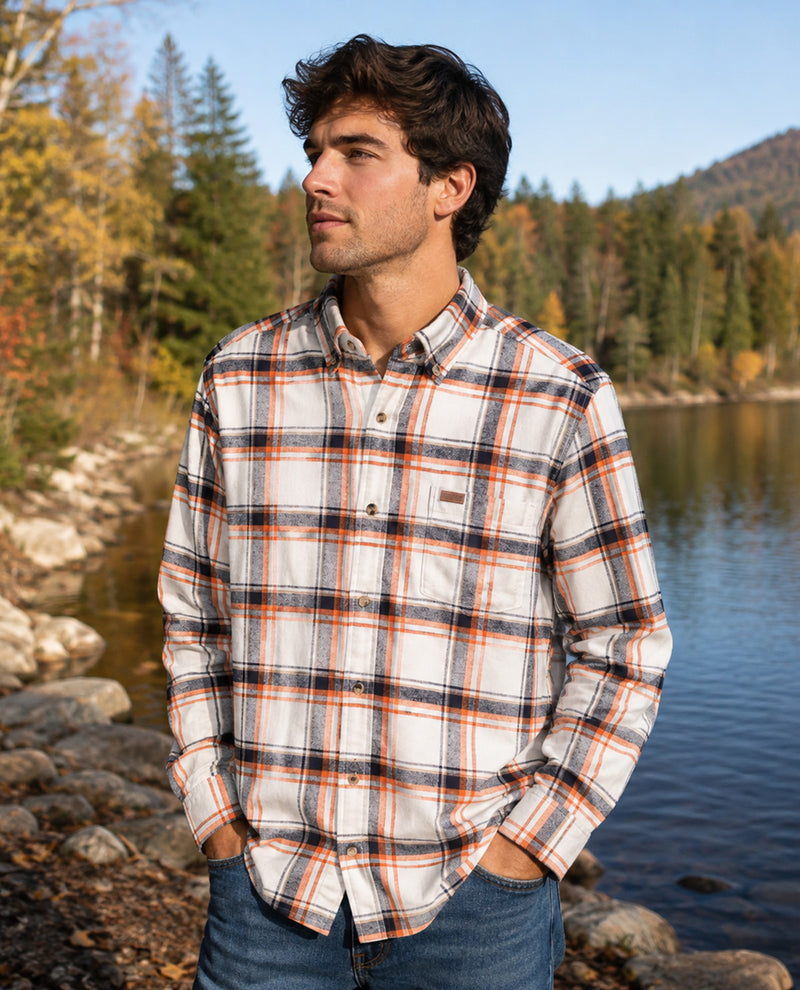 A man wearing a long-sleeve plaid button-down flannel shirt in white, orange, and black, standing outdoors by a lake with trees in the background.