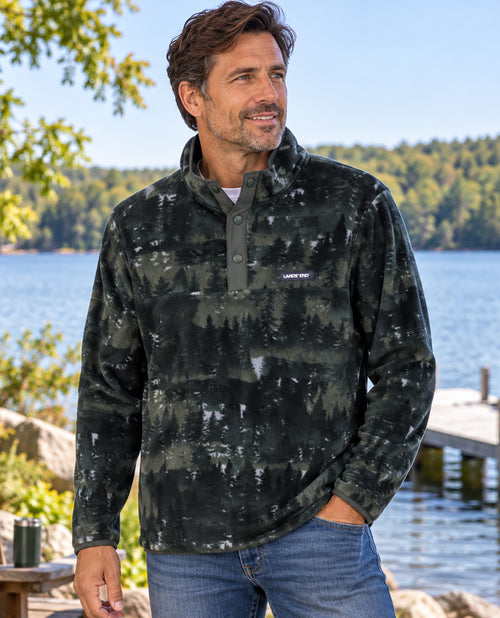 A man wearing a dark camouflage quarter-zip fleece pullover with blue jeans, standing outdoors near a lake with trees and a dock in the background.