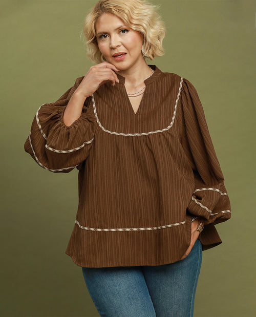 A woman wearing a brown long-sleeve blouse with cream-colored decorative piping and blue jeans, standing against a solid olive green background.