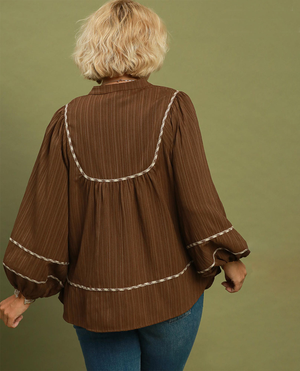 A woman viewed from the back wearing a brown long-sleeve blouse with cream-colored braided trim details, paired with blue jeans, set against a solid olive green background.