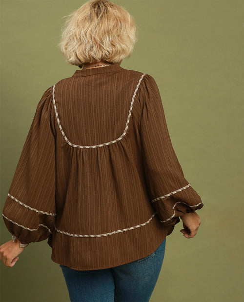 A woman viewed from the back wearing a brown long-sleeve blouse with cream-colored braided trim details, paired with blue jeans, set against a solid olive green background.