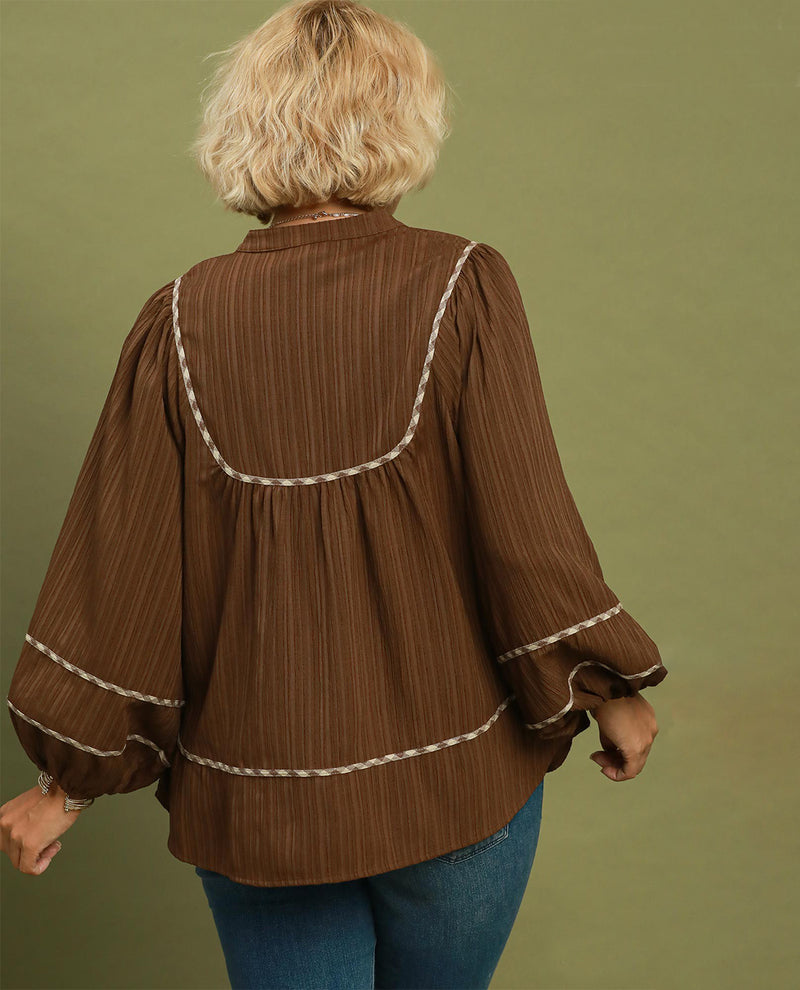 A woman viewed from the back wearing a brown long-sleeve blouse with cream-colored braided trim details, paired with blue jeans, set against a solid olive green background.