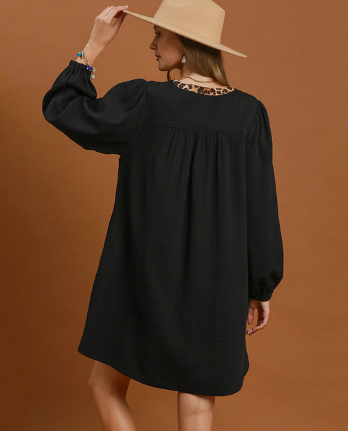 A woman wearing a black long-sleeve puff-sleeve dress with a leopard-print lace neckline, shown from the back against a solid brown studio background.