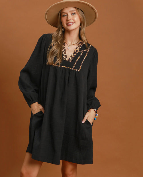 A woman wearing a loose black long-sleeve dress with a leopard-print embroidered neckline, styled with a wide-brimmed tan hat against a solid brown studio background.