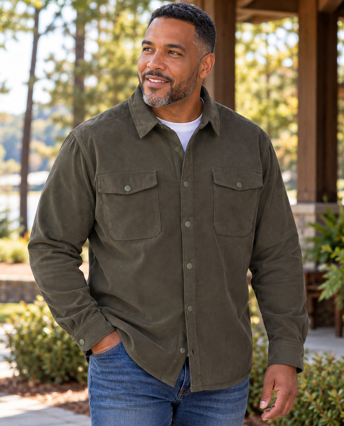 A man wearing an olive green corduroy button-up shirt with two chest pockets, paired with blue jeans, standing outdoors in front of a wooden covered walkway.