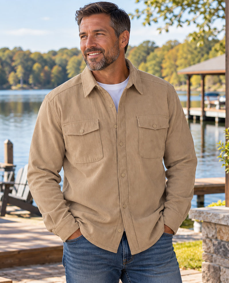 A man wearing a tan corduroy button-down shirt with two chest pockets over a white t-shirt and blue jeans, standing outdoors by a lake with a wooden dock and trees in the background.