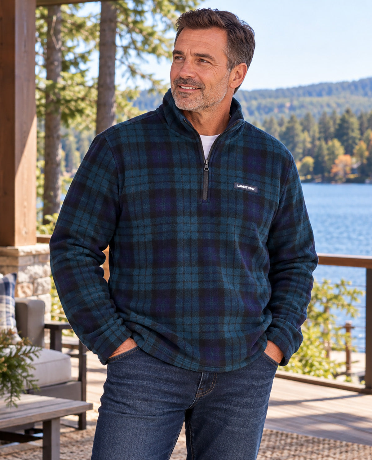 A man wearing a dark green and navy plaid quarter-zip fleece pullover jacket with a small visible Patagonia logo on the chest, standing on a wooden deck with a lake and forest in the background.