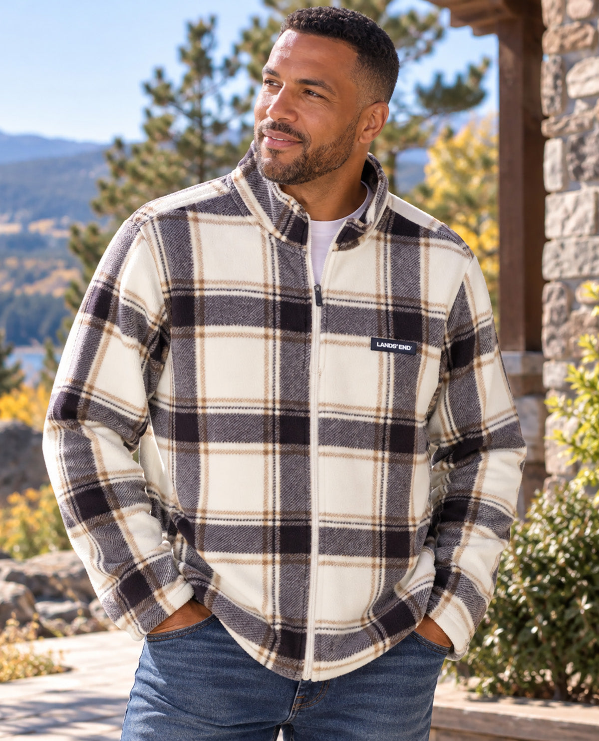 A man wearing a Patagonia fleece jacket with a brown, black, and cream plaid pattern, standing outdoors in front of a stone building and trees.