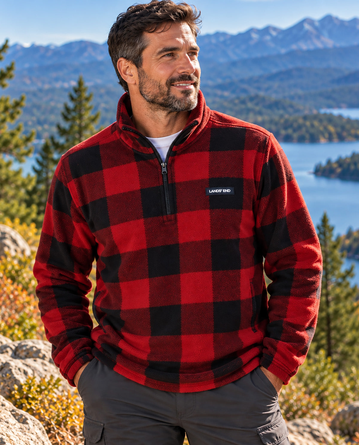 A man wearing a red and black buffalo plaid quarter-zip fleece pullover by Laux Pro, standing outdoors with a mountain and lake landscape in the background.