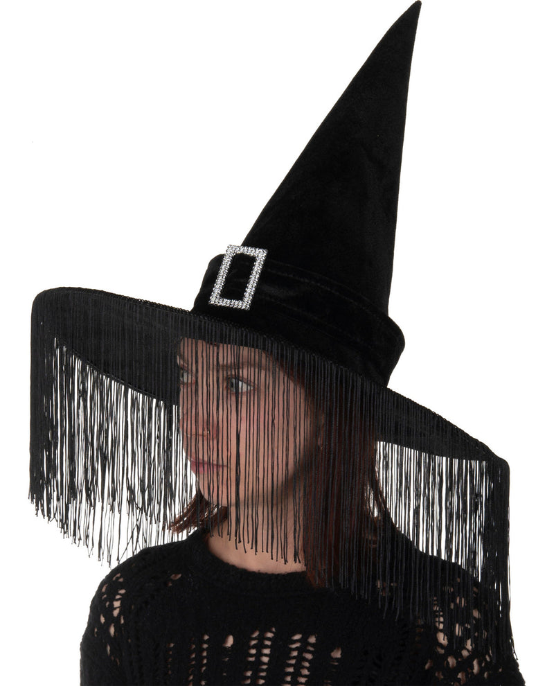 A black witch costume hat with a tall pointed crown, wide brim, and long fringe trim, worn by a model against a plain white background.