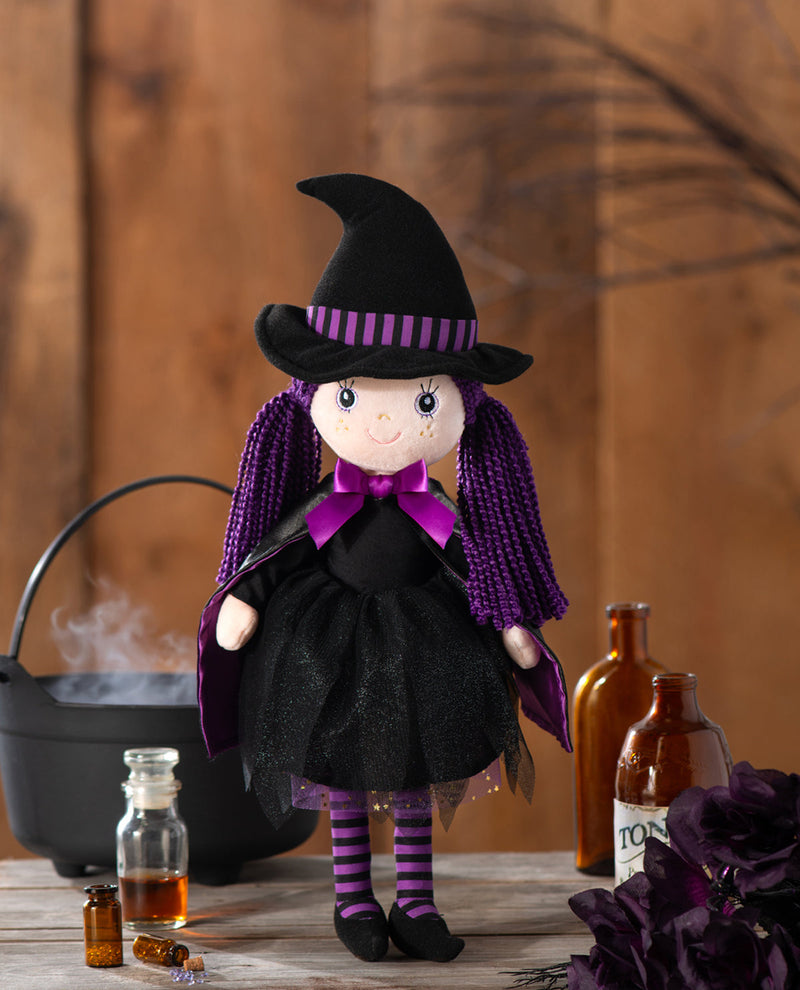 A witch doll with a black pointed hat, purple striped hatband, black dress, purple cape, and purple-and-black striped tights, posed on a rustic wooden surface with a cauldron, bottles, and flowers in a styled setting.