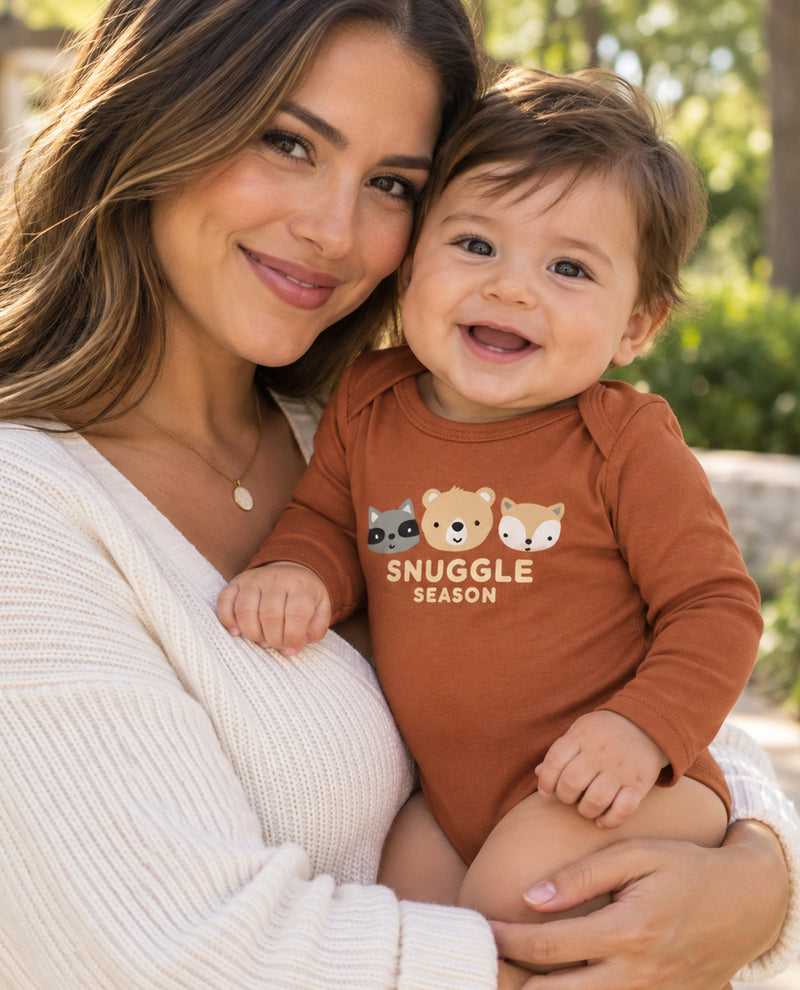 A smiling woman holding a baby outdoors, both wearing cozy knit clothing. The baby is wearing a rust-brown long-sleeve bodysuit with a graphic of cartoon animals and the text 'SNUGGLE SEASON'.