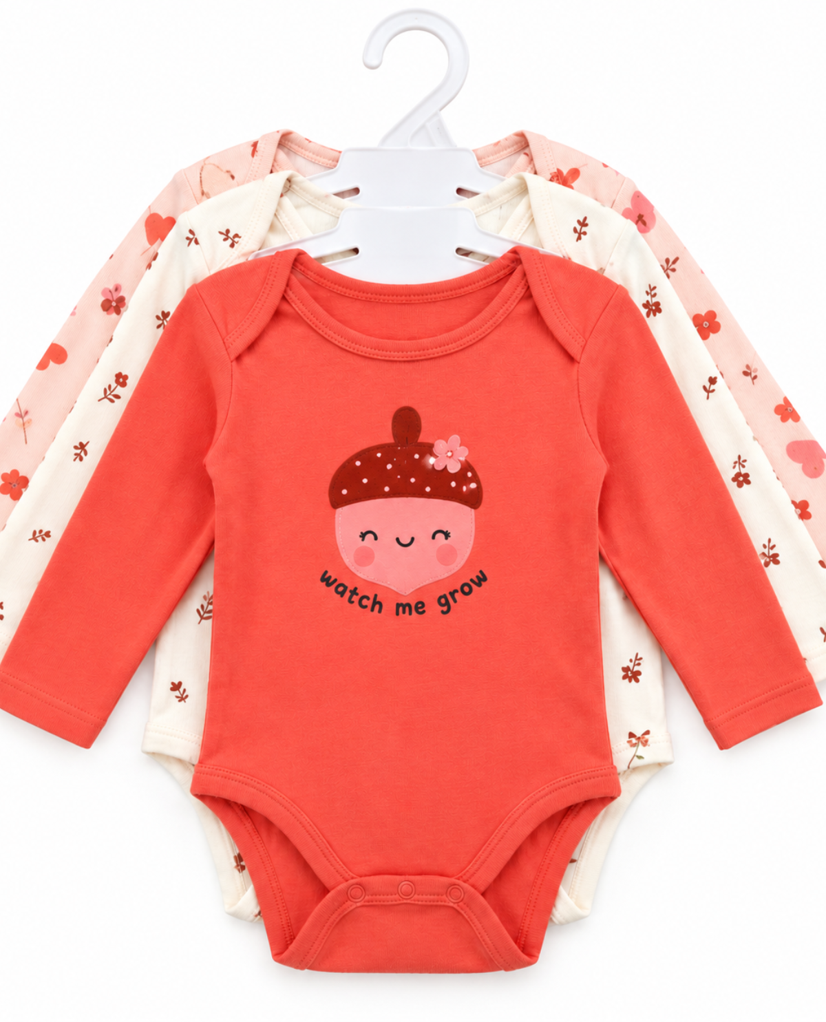 A set of three baby long-sleeve bodysuits hanging on a white plastic hanger against a white background. The front bodysuit is coral red with a cute smiling acorn graphic and the text 'watch me grow'.