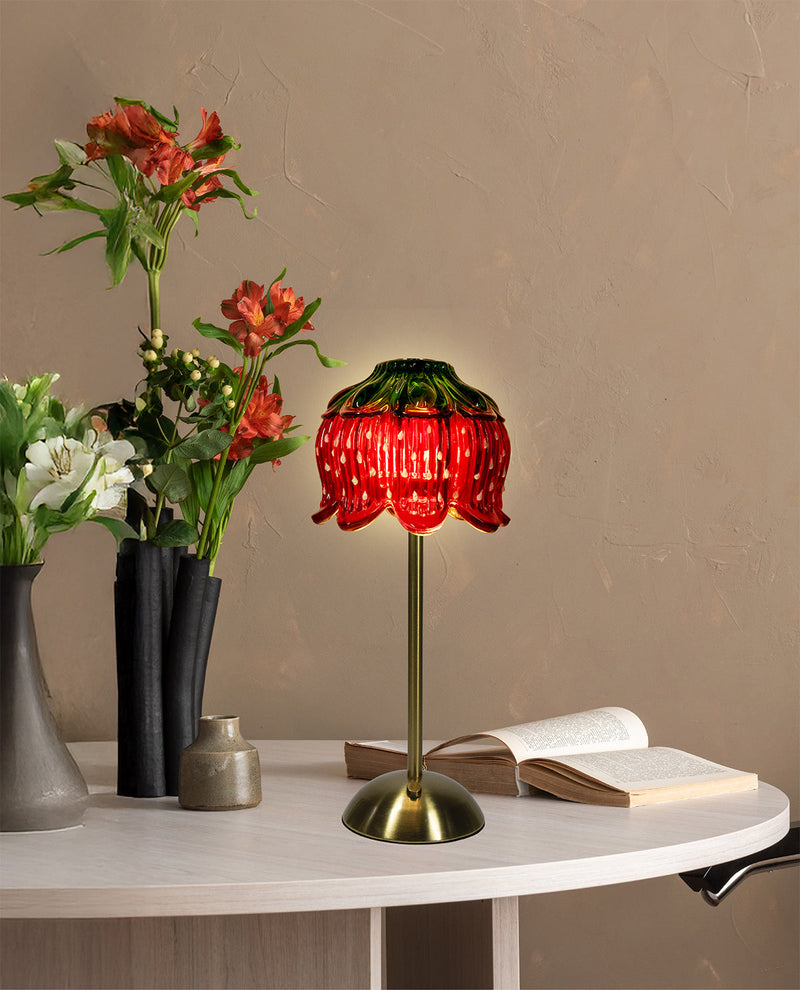 A red strawberry-shaped table lamp with a green top and gold base, styled on a white table next to vases of flowers and an open book against a beige wall.