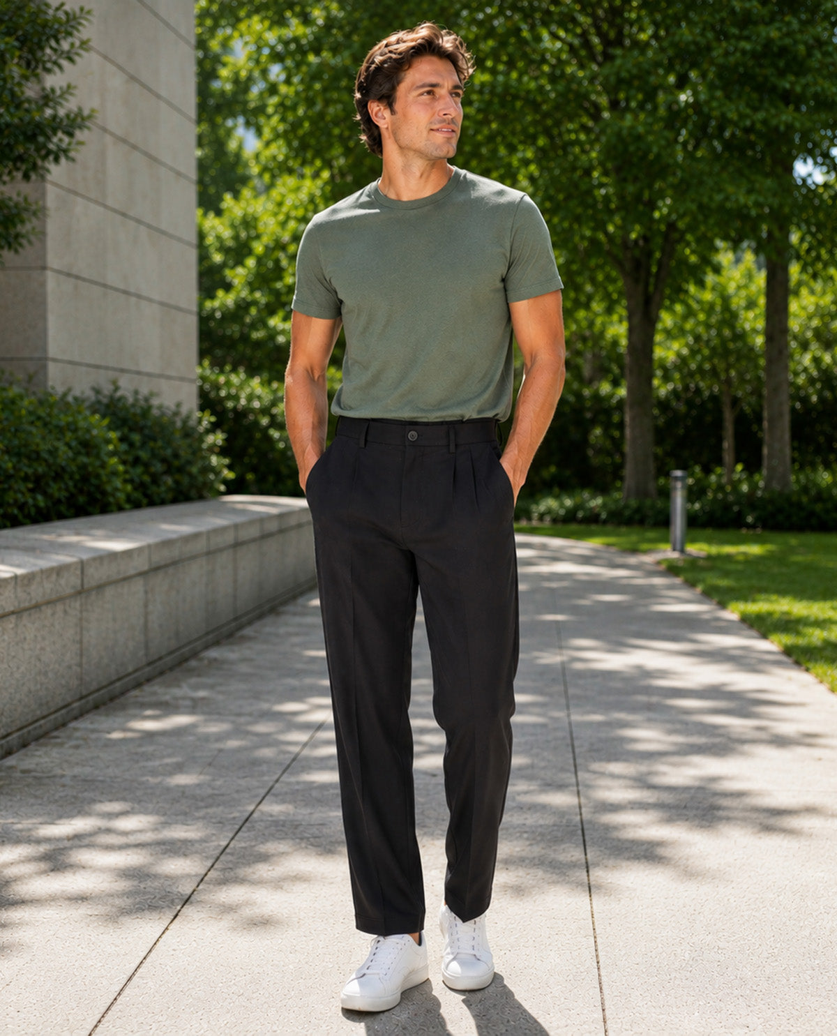 A man standing outdoors on a paved walkway wearing an olive green short-sleeve crewneck t-shirt, black pleated trousers, and white sneakers.